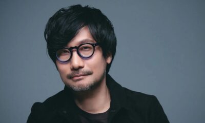 Hideo Kojima com óculos redondos, barba rala e cabelo escuro contra fundo azul-acinzentado