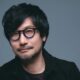 Hideo Kojima com óculos redondos, barba rala e cabelo escuro contra fundo azul-acinzentado