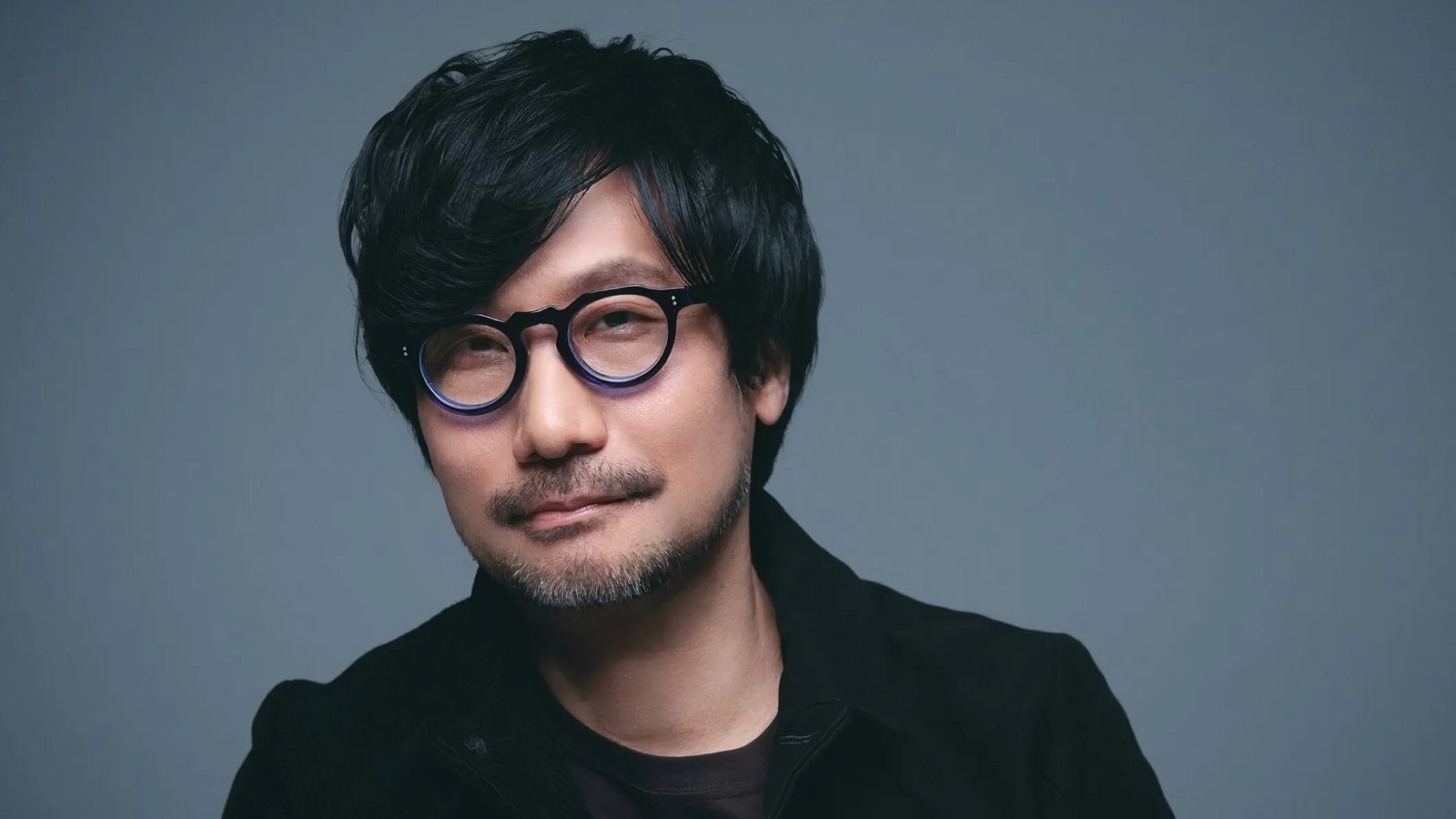 Hideo Kojima com óculos redondos, barba rala e cabelo escuro contra fundo azul-acinzentado