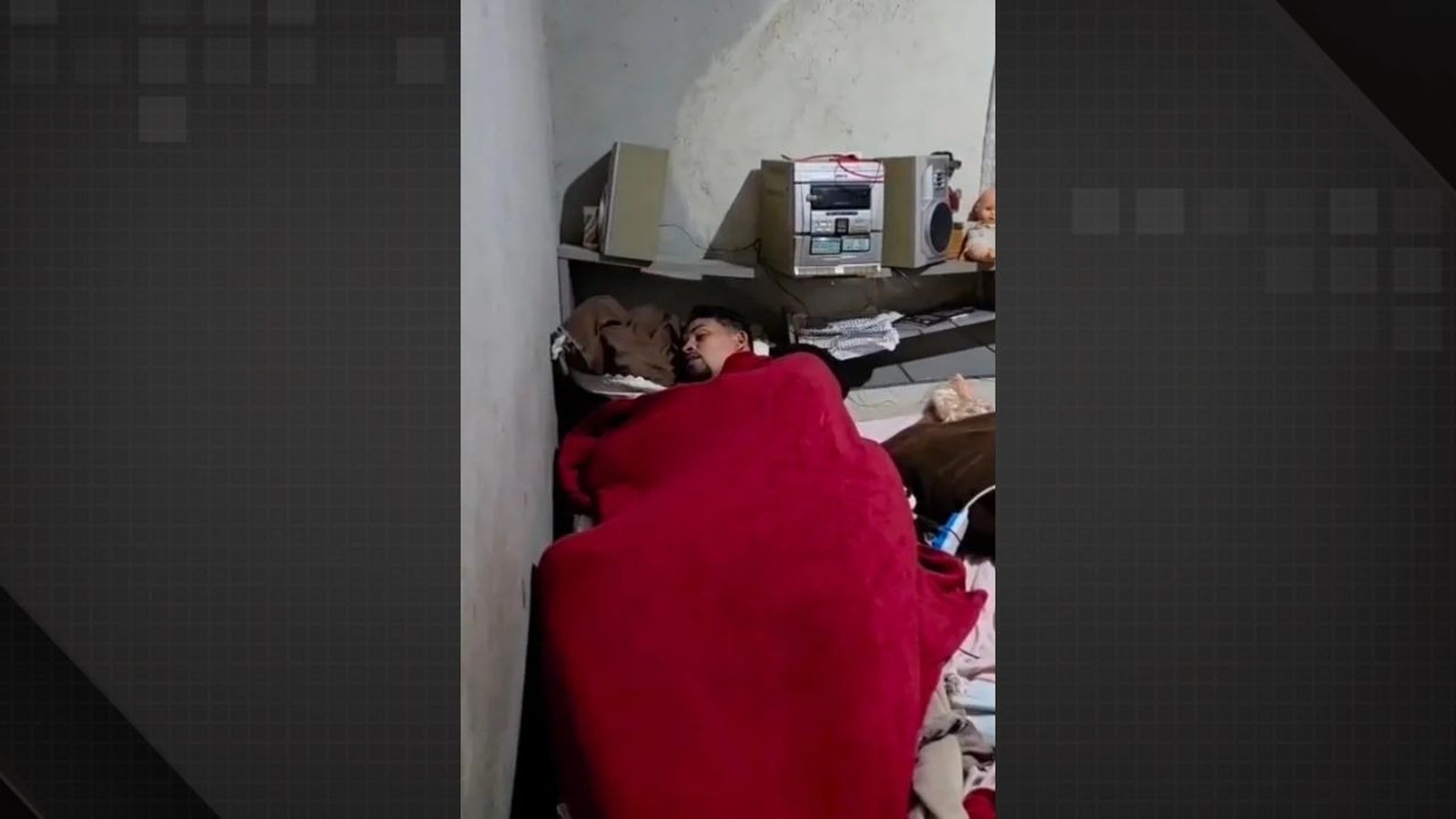 Homem deitado em cama, coberto por edredom vermelho, dormindo em quarto com aparelho de som