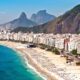 Praia urbana com mar azul, areia lotada de pessoas, edifícios e montanhas verdes
