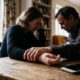 Um casal deprimido de cabeça baixa sentado à mesa de madeira com um smartphone e as mãos dadas.