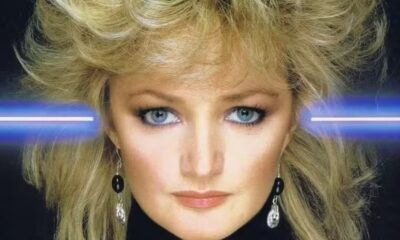 Close-up de Bonnie Tyler com cabelo loiro volumoso, olhos azuis, brincos e raios de luz azuis.
