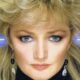Close-up de Bonnie Tyler com cabelo loiro volumoso, olhos azuis, brincos e raios de luz azuis.