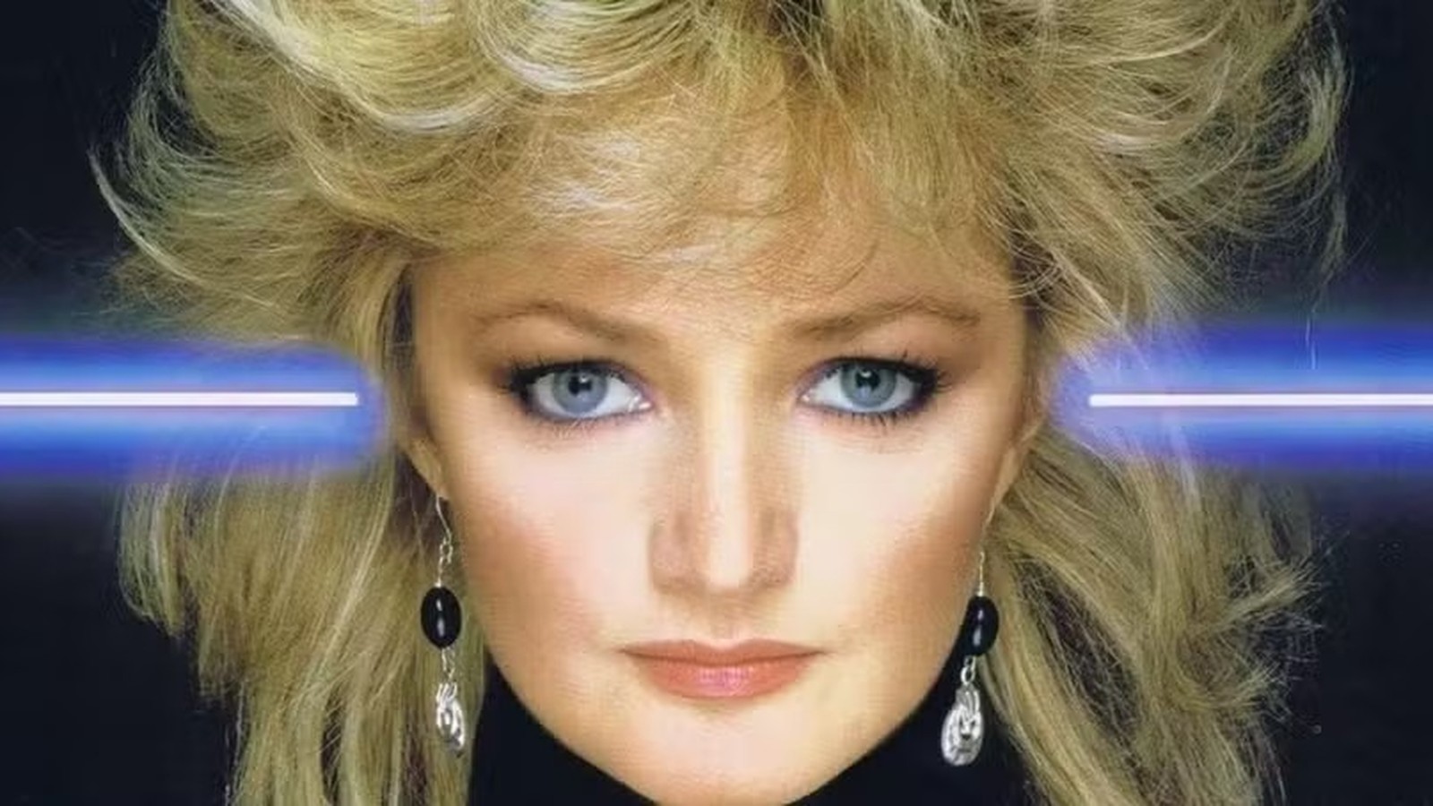 Close-up de Bonnie Tyler com cabelo loiro volumoso, olhos azuis, brincos e raios de luz azuis.