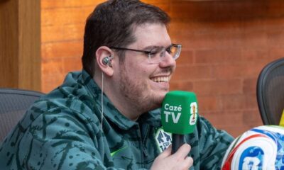 Casimiro sorrindo, usando óculos e fone, segurando microfone da CazéTV e uma bola de futebol