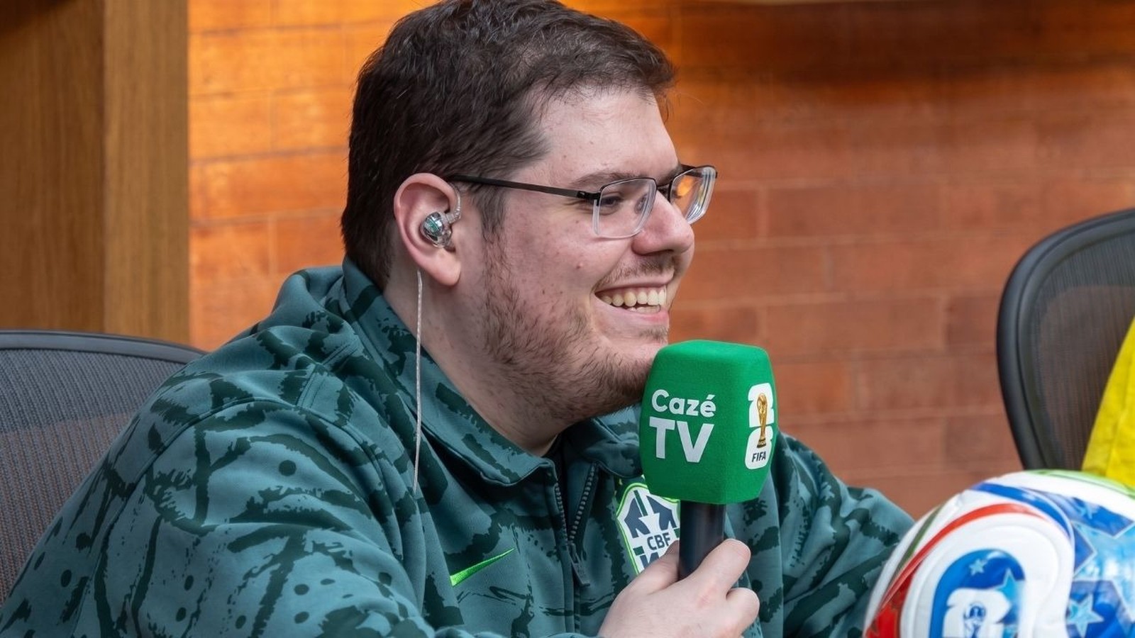 Casimiro sorrindo, usando óculos e fone, segurando microfone da CazéTV e uma bola de futebol