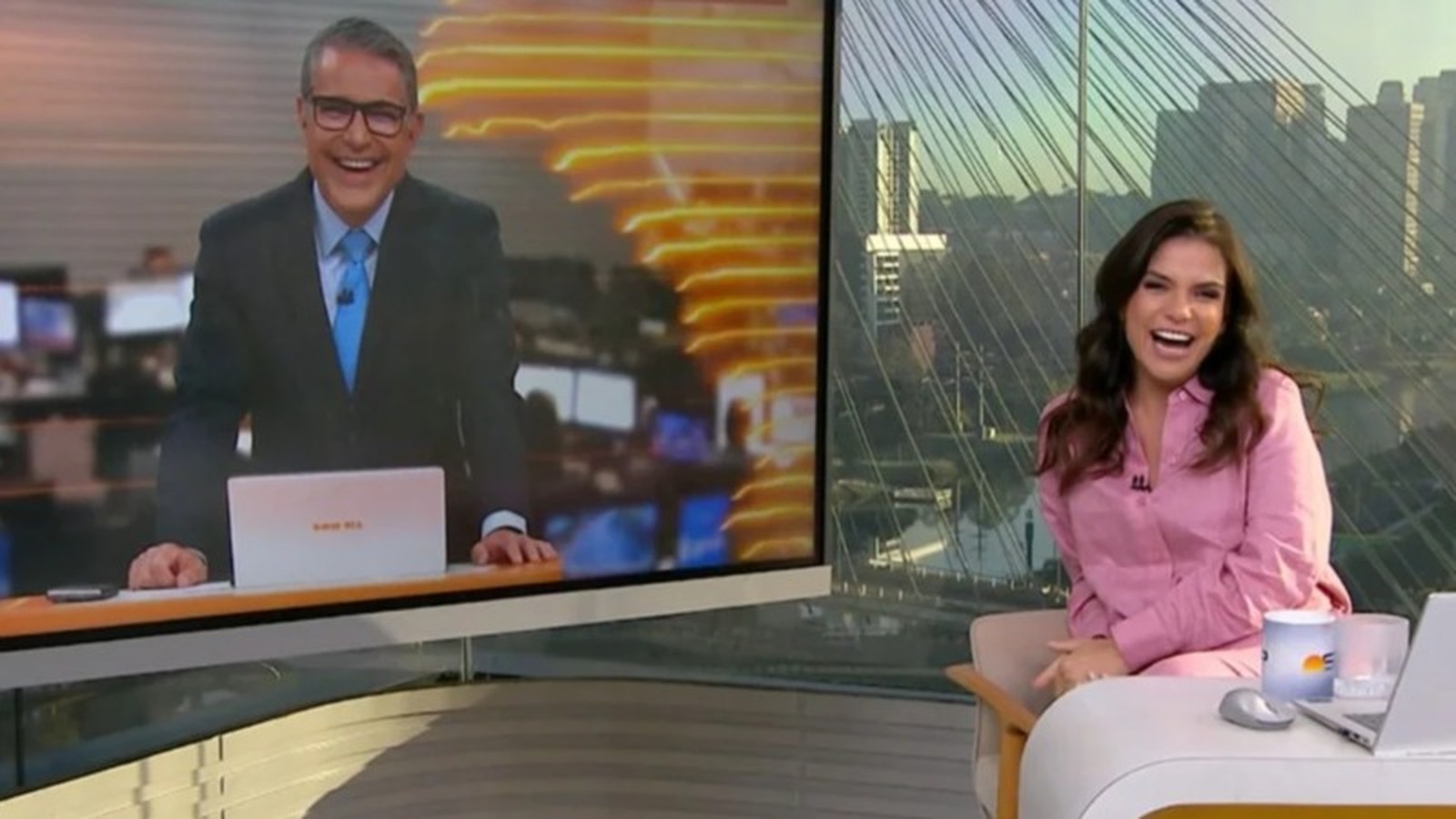 Hélter Duarte na tela e Sabina Simonato rindo em estúdio, com laptop e vista urbana