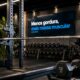 Academia moderna com barra carregada no suporte, halter ao lado e painel digital escrito “Menos gordura, mais massa muscular