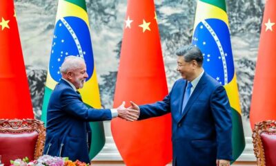 Lula e Xi Jinping sorrindo apertam as mãos com bandeiras do Brasil e China ao fundo