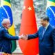Lula e Xi Jinping sorrindo apertam as mãos com bandeiras do Brasil e China ao fundo