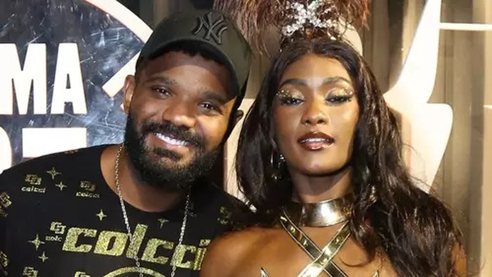 Erika Januza e Arlindinho sorrindo, ele de boné, ela com maquiagem dourada e roupa de festa