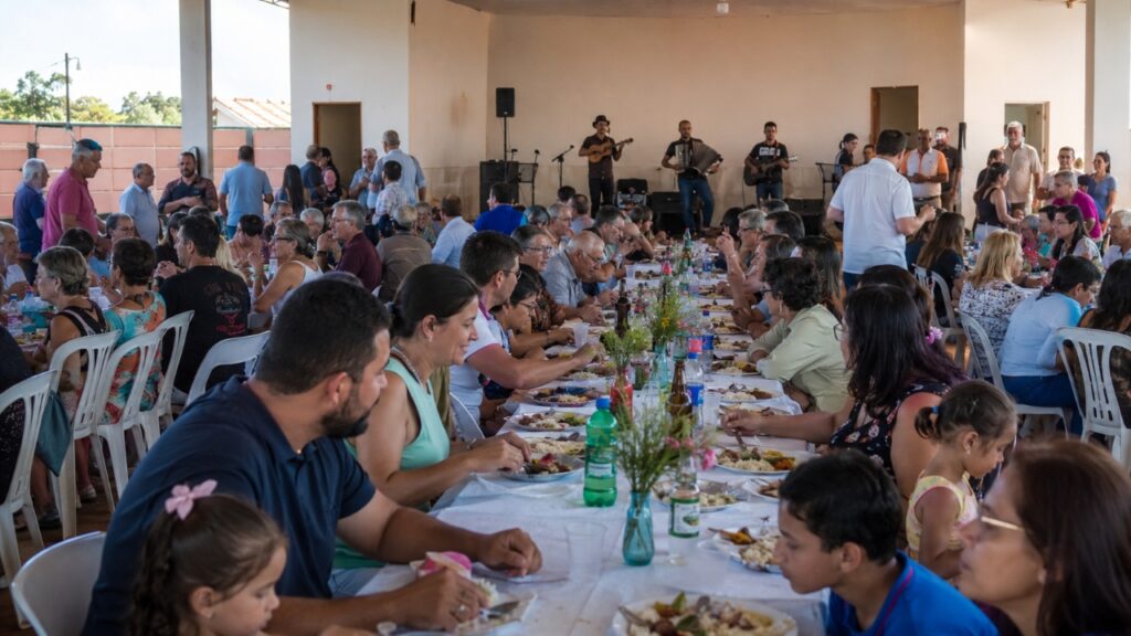 Pessoas reunidas em mesas durante o Almoço dos Passos, com pratos servidos e apresentação musical ao fundo em espaço comunitário de Rio Preto, Minas Gerais.