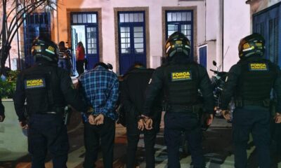 Dois agentes da Força Municipal com dois homens algemados vistos de costas à noite