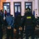 Dois agentes da Força Municipal com dois homens algemados vistos de costas à noite