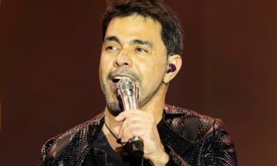 Zezé Di Camargo canta ao microfone no palco, usando fone de ouvido roxo.