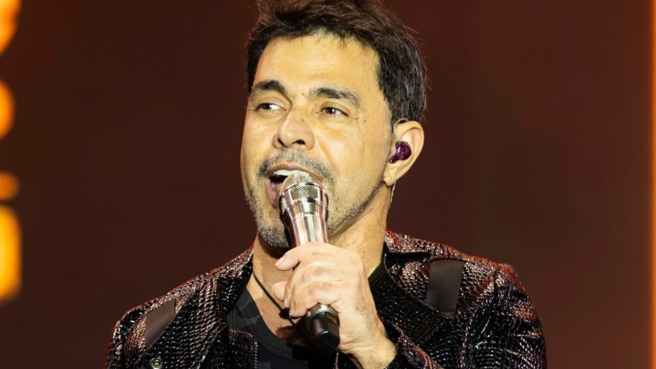 Zezé Di Camargo canta ao microfone no palco, usando fone de ouvido roxo.