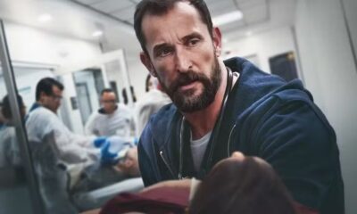 Médico barbudo com estetoscópio em hospital, olhando fixamente para frente; equipe médica ao fundo