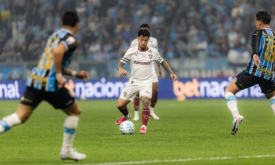 Jogador com camisa branca e grená dribla bola em campo, outros jogadores e torcida ao fundo