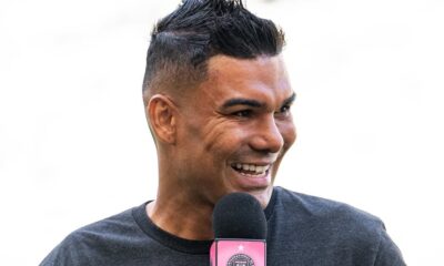 Homem sorrindo, cabelo espetado, camiseta cinza, falando em microfone com base rosa e logo