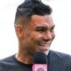 Homem sorrindo, cabelo espetado, camiseta cinza, falando em microfone com base rosa e logo
