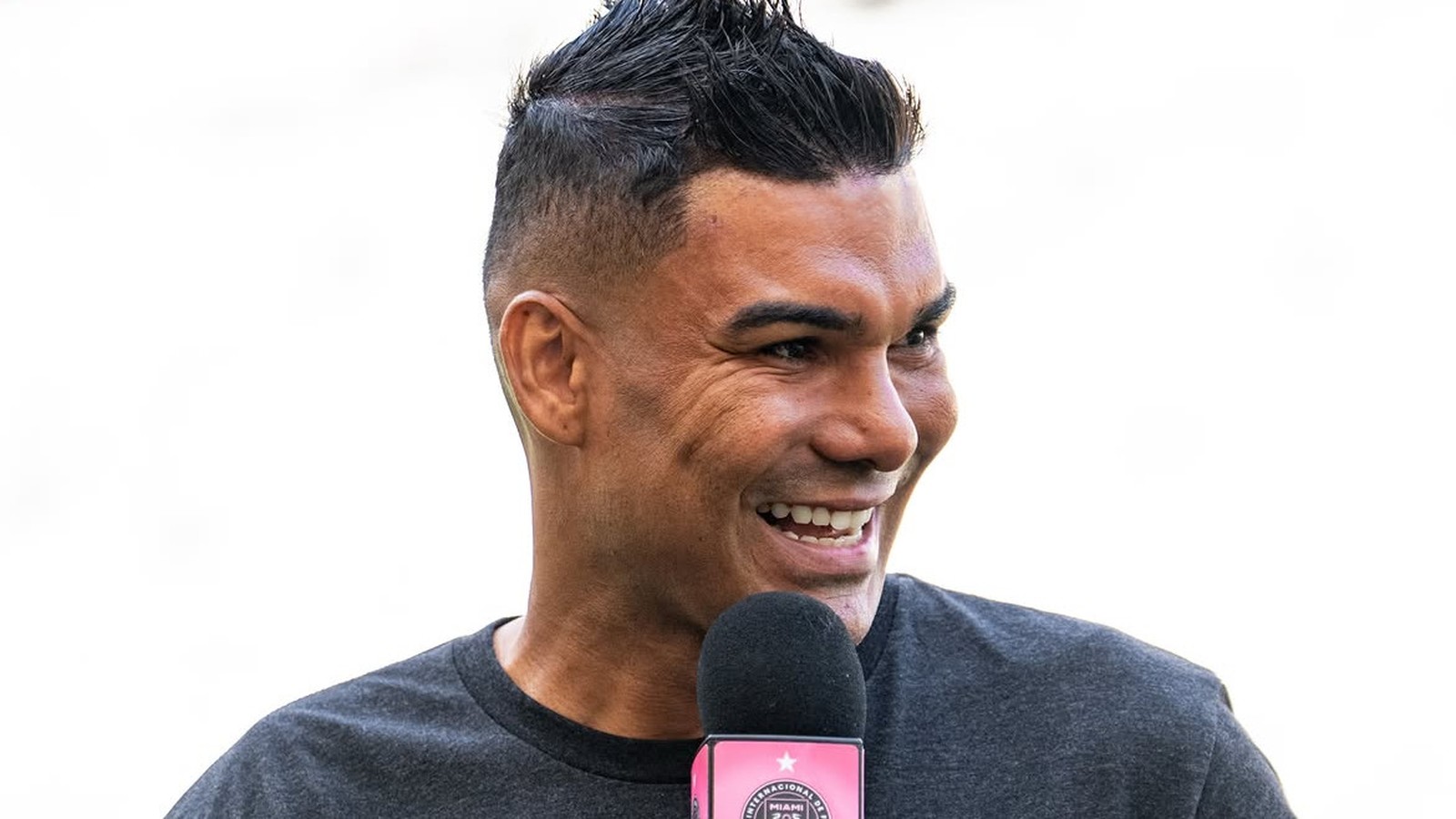 Homem sorrindo, cabelo espetado, camiseta cinza, falando em microfone com base rosa e logo