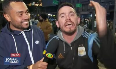 Repórter sorrindo segura microfone da Band e torcedor gesticula. Texto 20:00 Jornal da Band