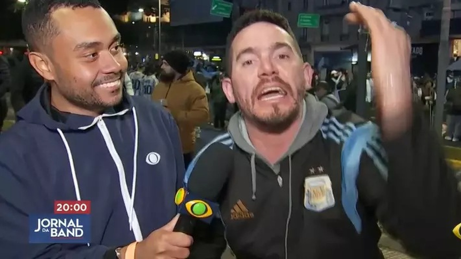 Repórter sorrindo segura microfone da Band e torcedor gesticula. Texto 20:00 Jornal da Band