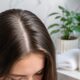 Mulher com cabelos castanhos e raiz limpa em banheiro moderno, com frasco de shampoo branco ao lado.