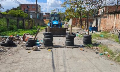 Barricada de pneus empilhados em rua, com retroescavadeira azul ao fundo e lixo