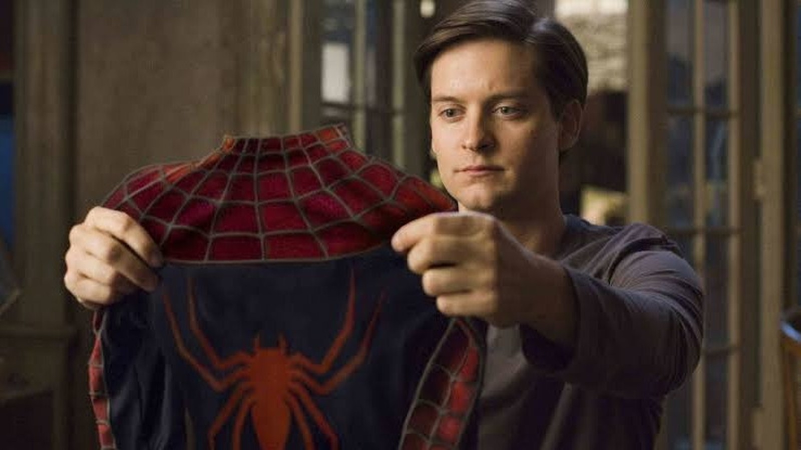 Tobey Maguire segura o traje do Homem-Aranha vermelho e azul, olhando para ele