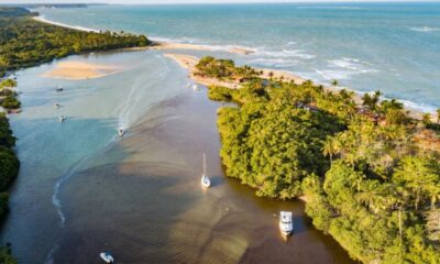 Vista aérea de rio com barcos, densa vegetação, unindo-se ao mar e praia arenosa