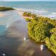 Vista aérea de rio com barcos, densa vegetação, unindo-se ao mar e praia arenosa