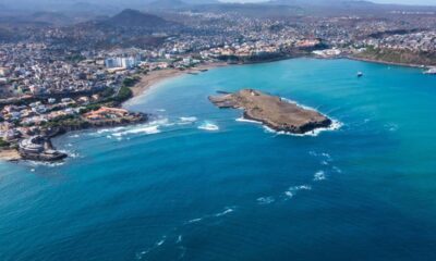 Vista aérea de cidade costeira em Cabo Verde com oceano azul, ilha rochosa e montanhas.