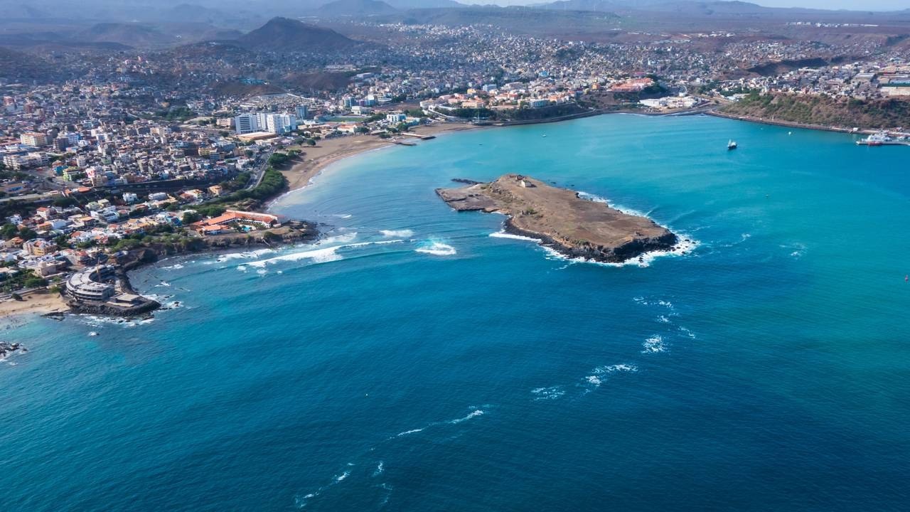 Vista aérea de cidade costeira em Cabo Verde com oceano azul, ilha rochosa e montanhas.