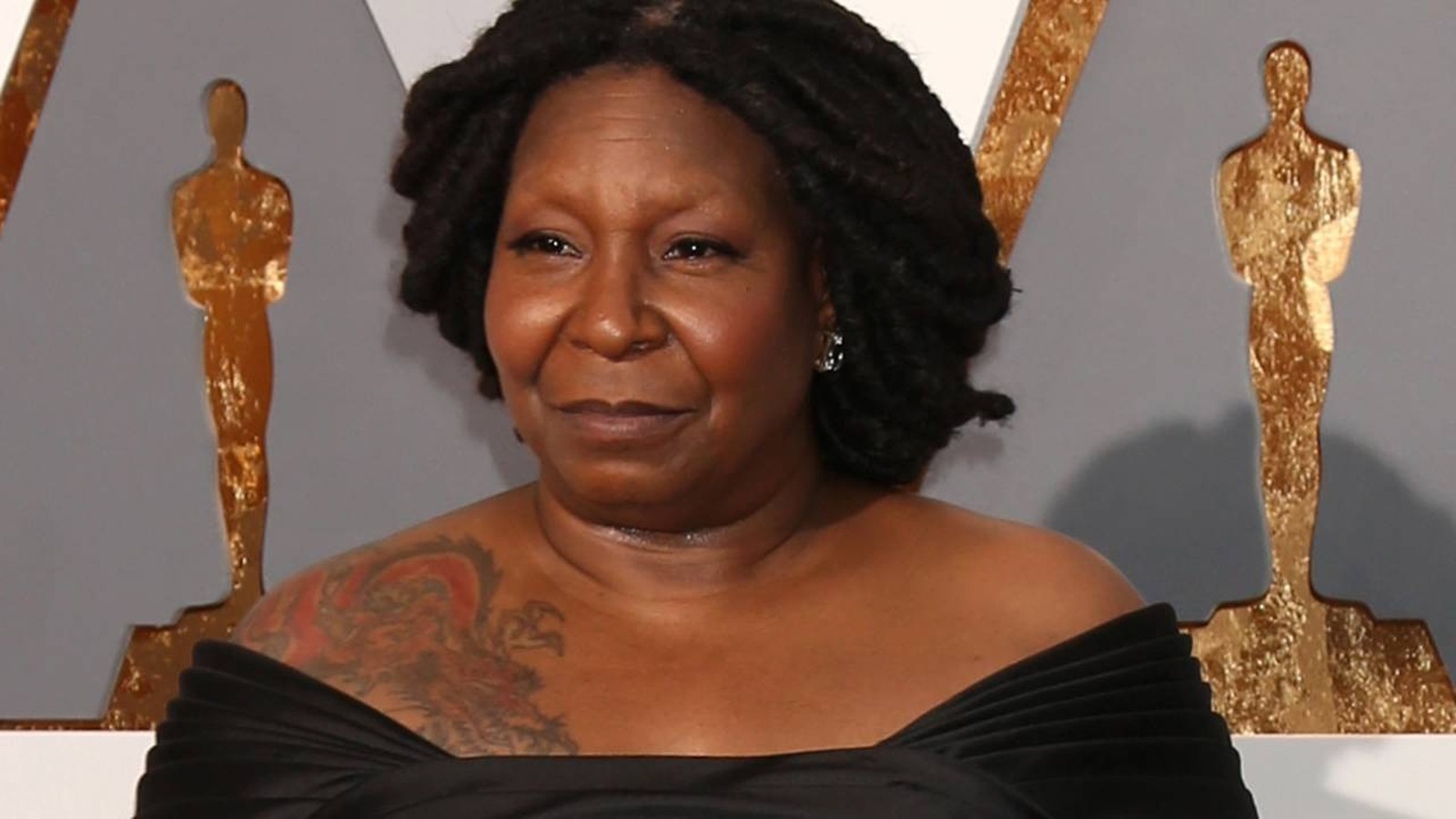Whoopi Goldberg, dreadlocks, vestido preto, tatuagem, fundo com silhuetas douradas de Oscar