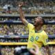 Jogador da seleção brasileira de camisa amarela celebra com braço erguido em estádio lotado.