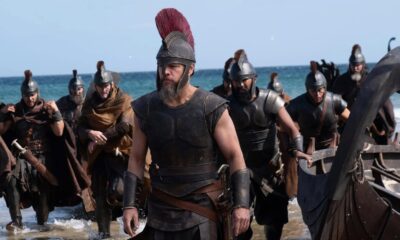Matt Damon como guerreiro grego, capacete e barba, saindo do mar com soldados e barco