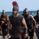 Matt Damon como guerreiro grego, capacete e barba, saindo do mar com soldados e barco