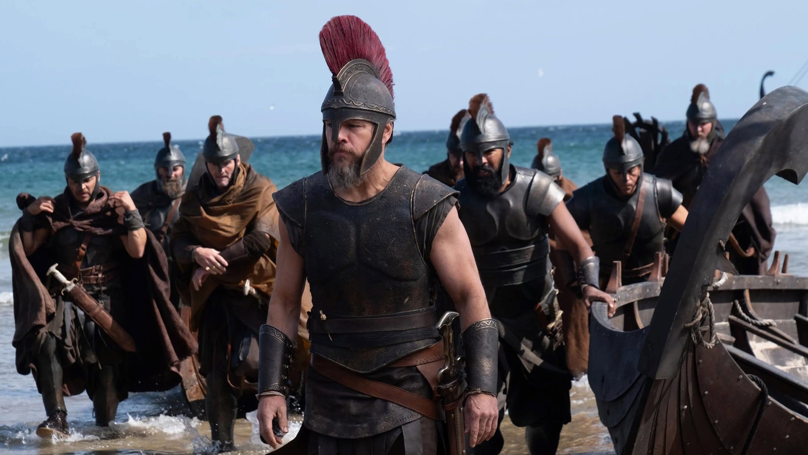 Matt Damon como guerreiro grego, capacete e barba, saindo do mar com soldados e barco