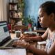 Mulher jovem concentrada estudando em laptop com caderno e caneta em mesa de madeira.