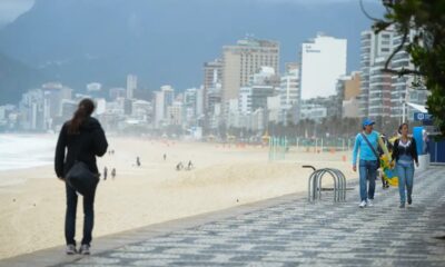 Vista da orla da praia com pessoas caminhando em calçadão, prédios e montanha ao fundo em dia nublado.