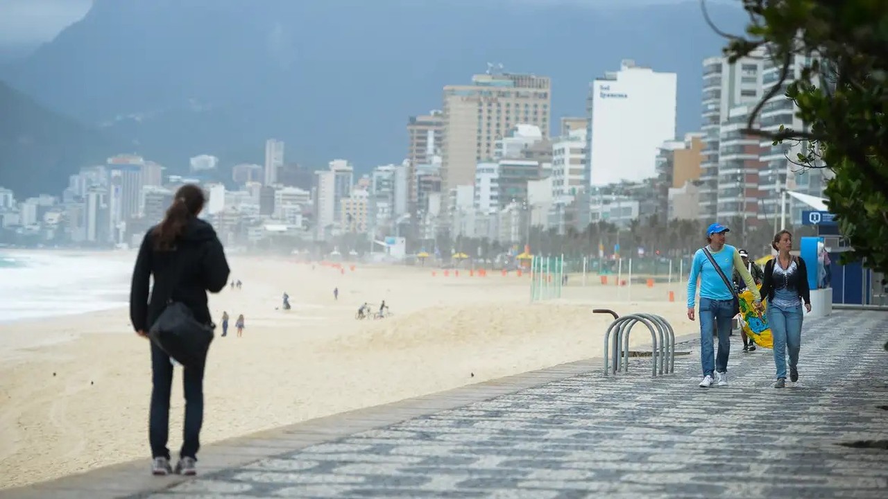 Vista da orla da praia com pessoas caminhando em calçadão, prédios e montanha ao fundo em dia nublado.
