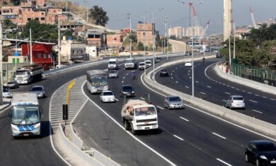 Rodovia movimentada com ônibus, carros e caminhões, e casas em morros ao fundo sob céu azul