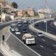 Rodovia movimentada com ônibus, carros e caminhões, e casas em morros ao fundo sob céu azul
