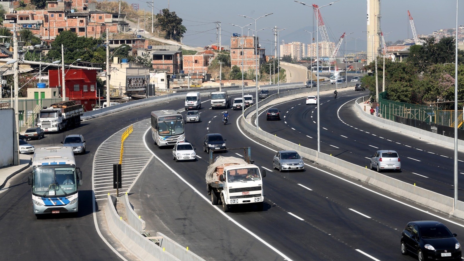 Rodovia movimentada com ônibus, carros e caminhões, e casas em morros ao fundo sob céu azul