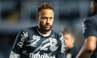 Neymar de barba e cabelo encaracolado, vestindo camisa Umbro do Santos FC com patrocinadores