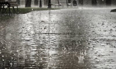Chuva intensa caindo sobre piso de paralelepípedos, com respingos visíveis e bancos desfocados