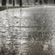 Chuva intensa caindo sobre piso de paralelepípedos, com respingos visíveis e bancos desfocados