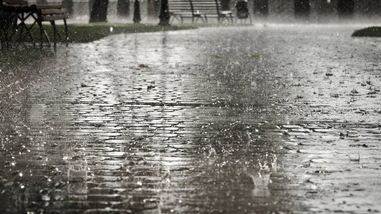 Chuva intensa caindo sobre piso de paralelepípedos, com respingos visíveis e bancos desfocados
